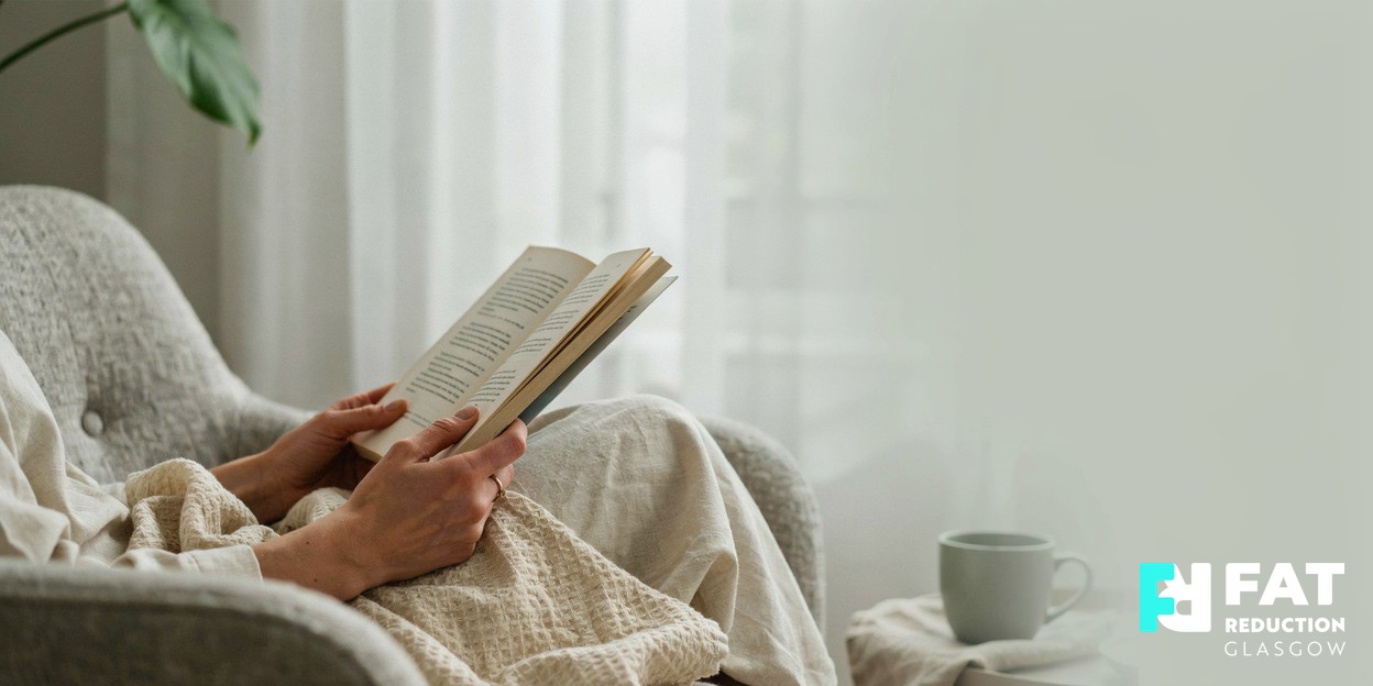 A person reclining comfortably in a soft grey clinic chair reading a book, wrapped in a light blanket, in warm natural daylight