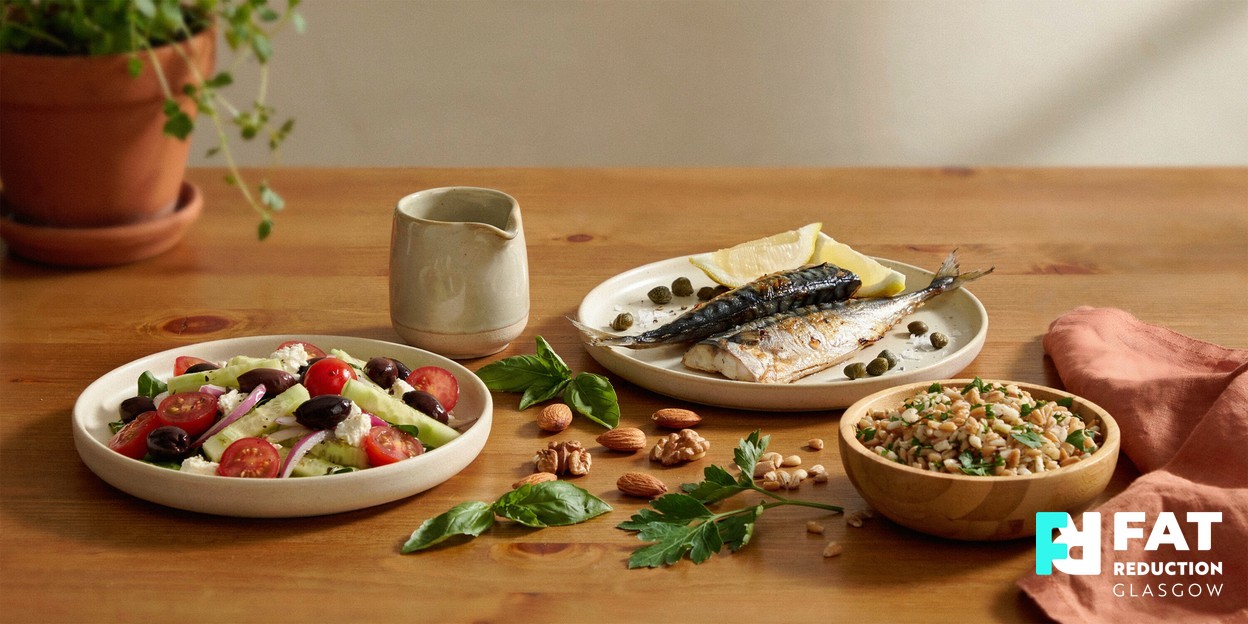 A wooden table with a fresh Mediterranean-style spread of olive oil, oily fish, wholegrains and colourful vegetables in warm daylight